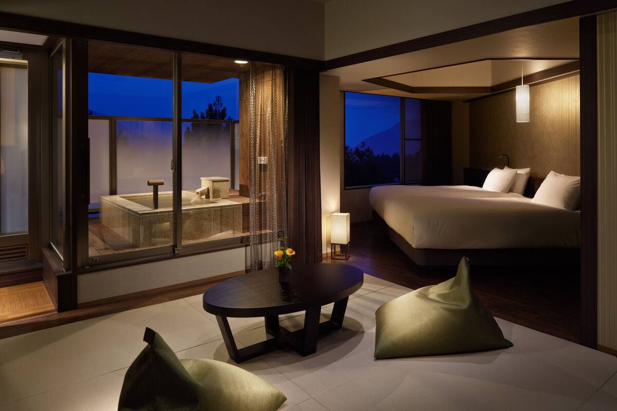 A modern hotel room with a large bed, a dining table, and a view of the outdoors.