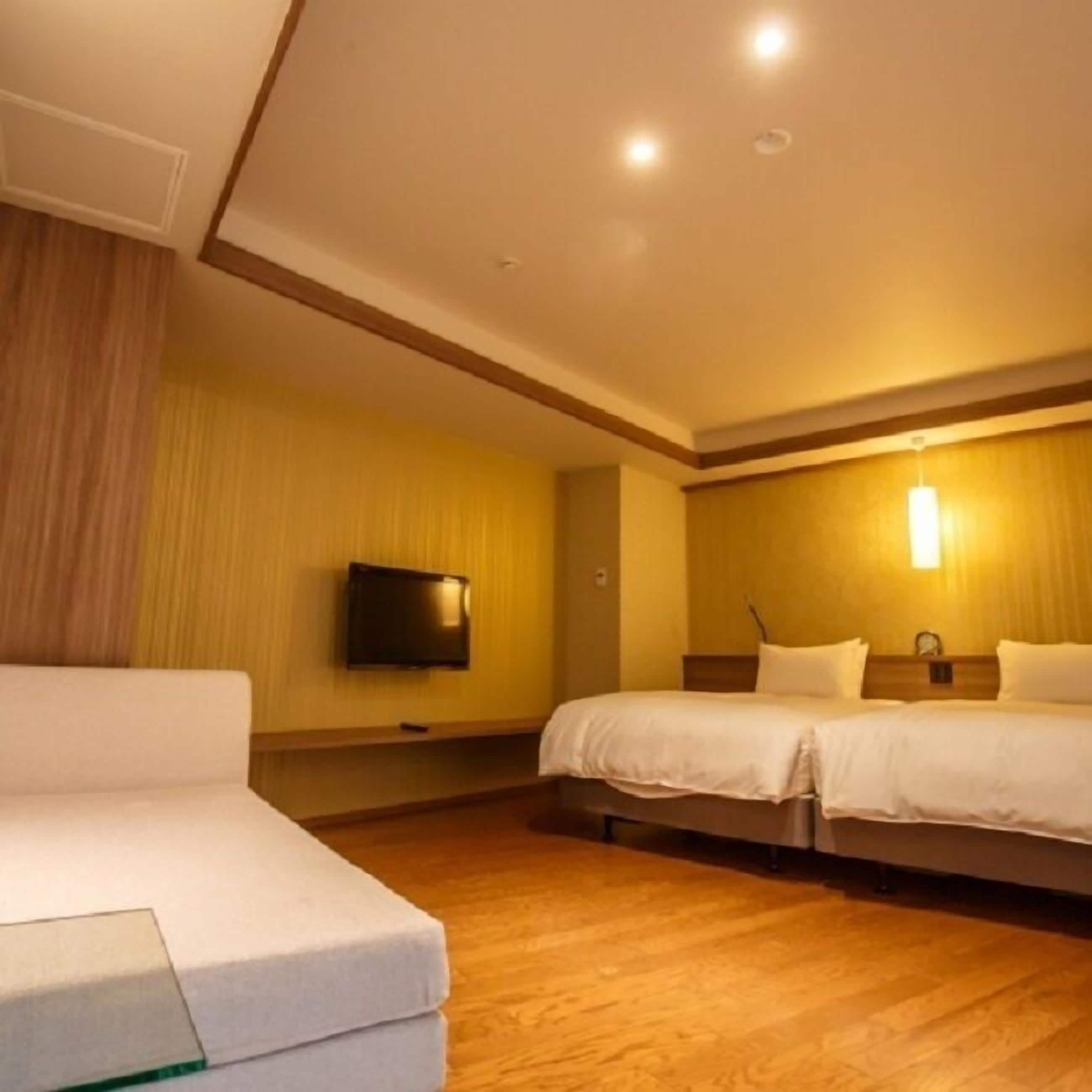 A hotel room with a sofa, two beds, a flat-screen TV, and a wooden floor.