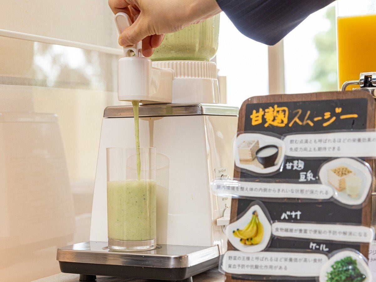 A person pouring a green liquid into a glass using a dispenser. A menu board with Japanese text is visible to the right.