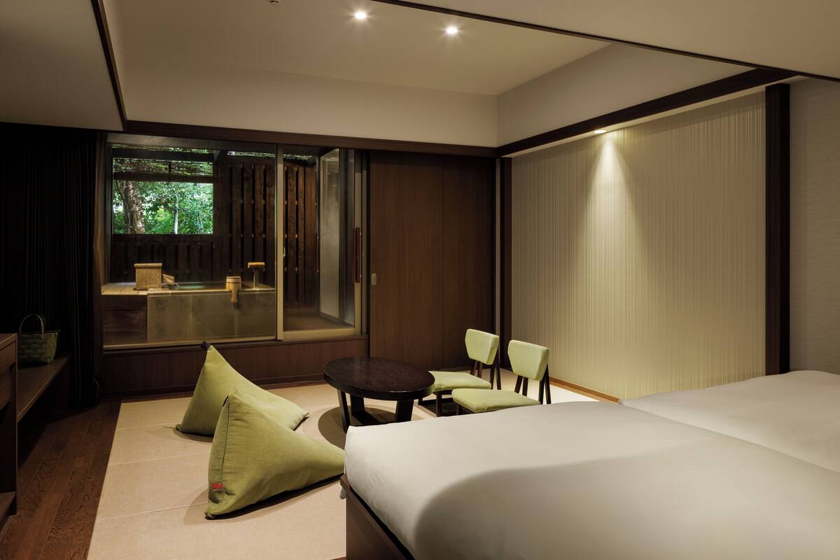 A modern hotel room with a large bed, a small round table, two chairs, and a large window offering a view of the outdoors.