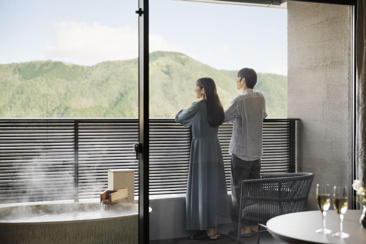 A couple standing by a window overlooking a mountainous landscape, with a table and chairs in the foreground.