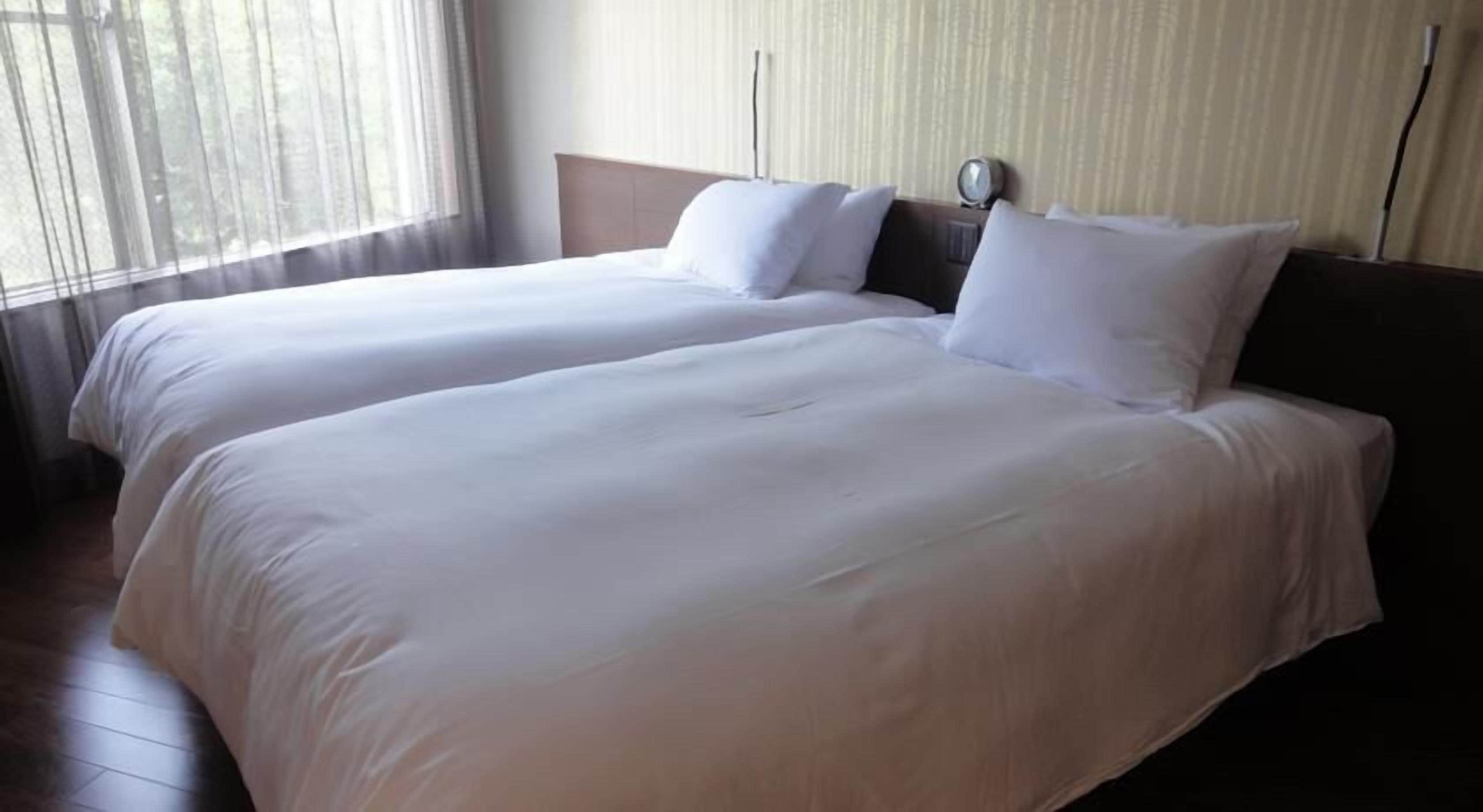 A hotel room with two single beds, a wooden headboard, a bedside table with a lamp, and a window with sheer curtains.
