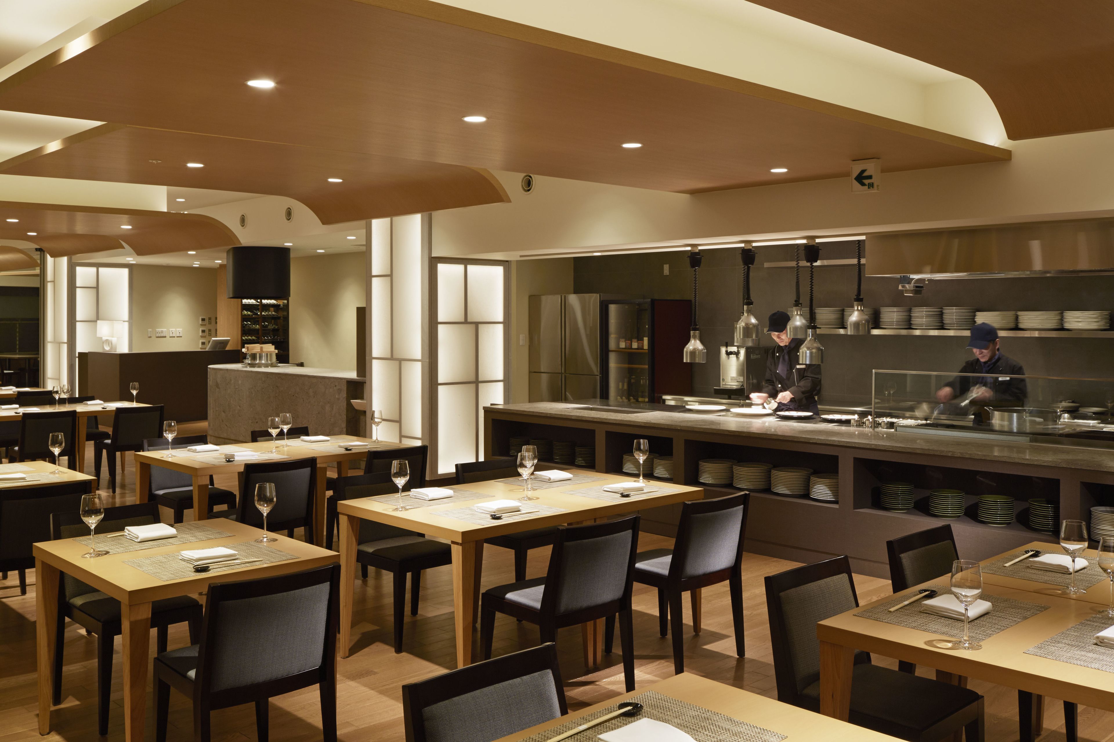 Een modern restaurant met houten tafels en stoelen, een centraal gelegen keuken en hangende verlichting.