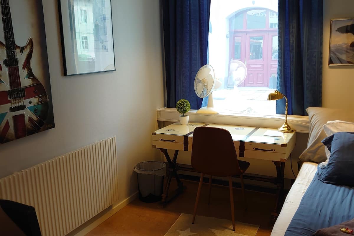 A bedroom with a bed, a desk with a chair, a radiator, a window with curtains, and a wall-mounted fan.