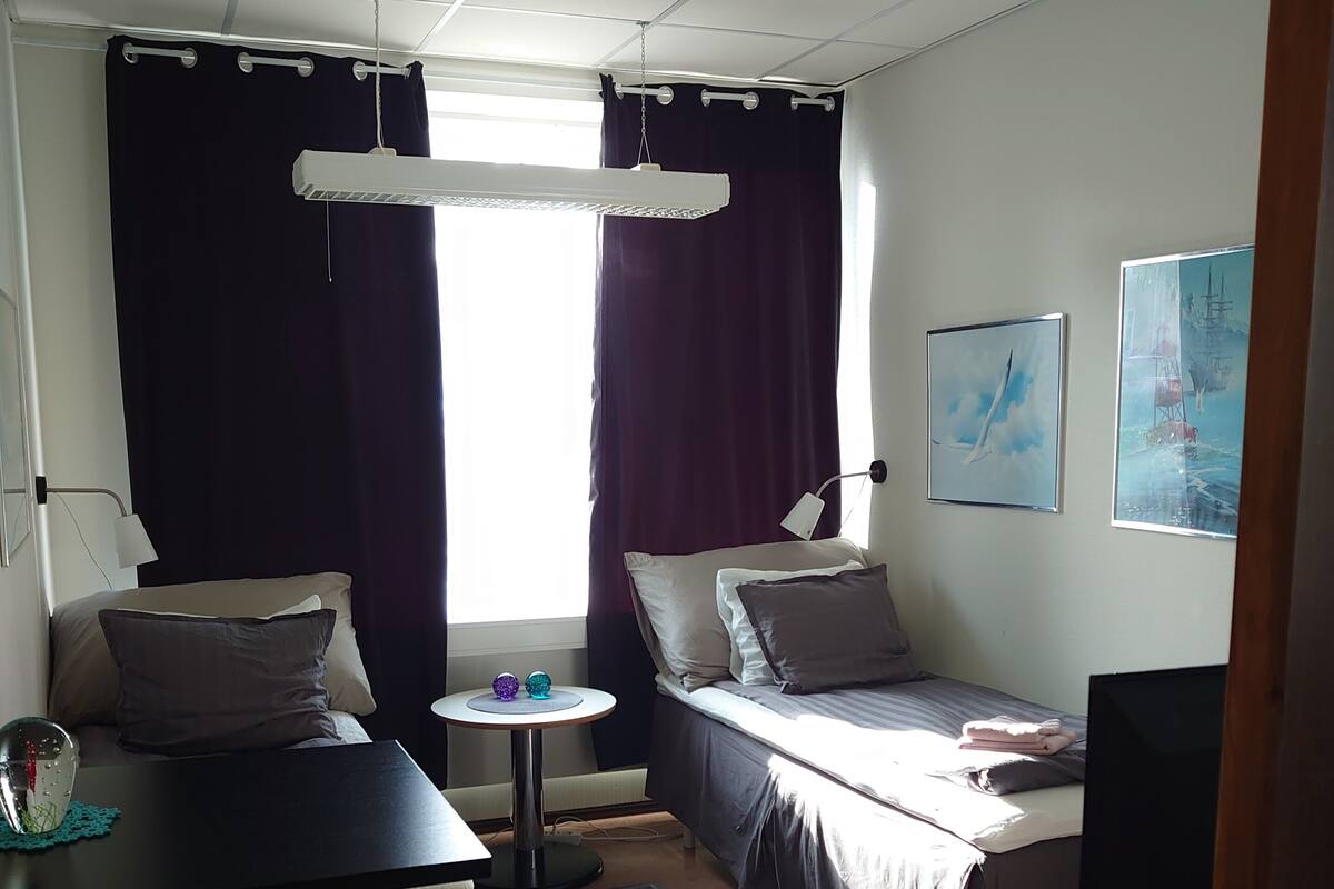 A compact hotel room with a bed, a desk, a chair, a small table, a TV, and a window with curtains.