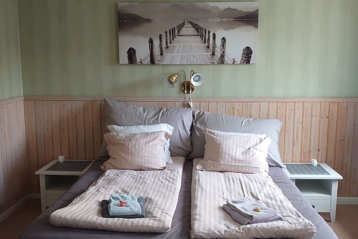 A double bed with a grey bedspread, two bedside tables with lamps, a wall-mounted painting of a pier, and a wooden headboard.