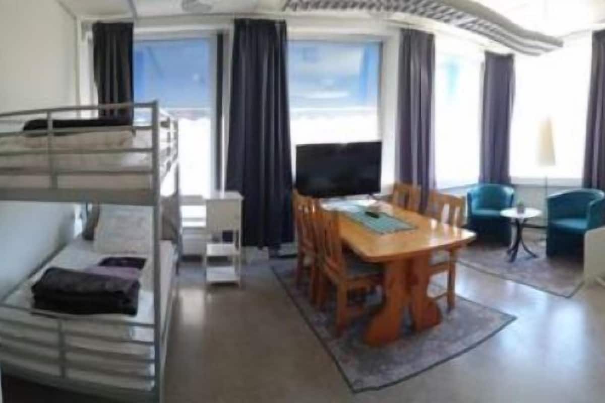A hotel room with a bunk bed, a dining table with chairs, a TV, a small round table, a sofa, a dresser, and a window with curtains.