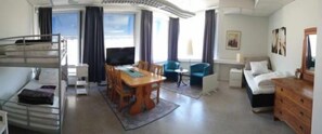 Une chambre d’hôtel avec un lit superposé, une table à manger avec des chaises, une télévision, une petite table ronde, un canapé, une commode et une fenêtre avec des rideaux.
