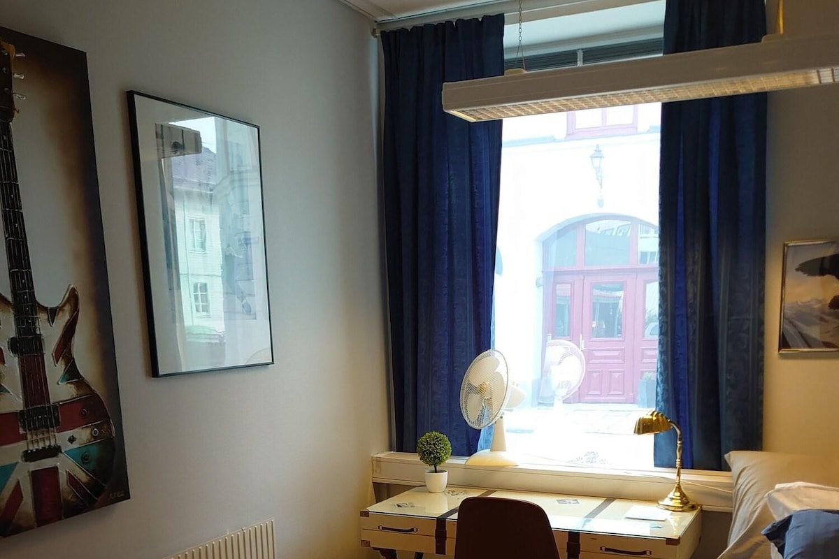 Una habitación con una cama, un escritorio con una silla, un radiador, una ventana con cortinas, un ventilador de pared y un cuadro enmarco.