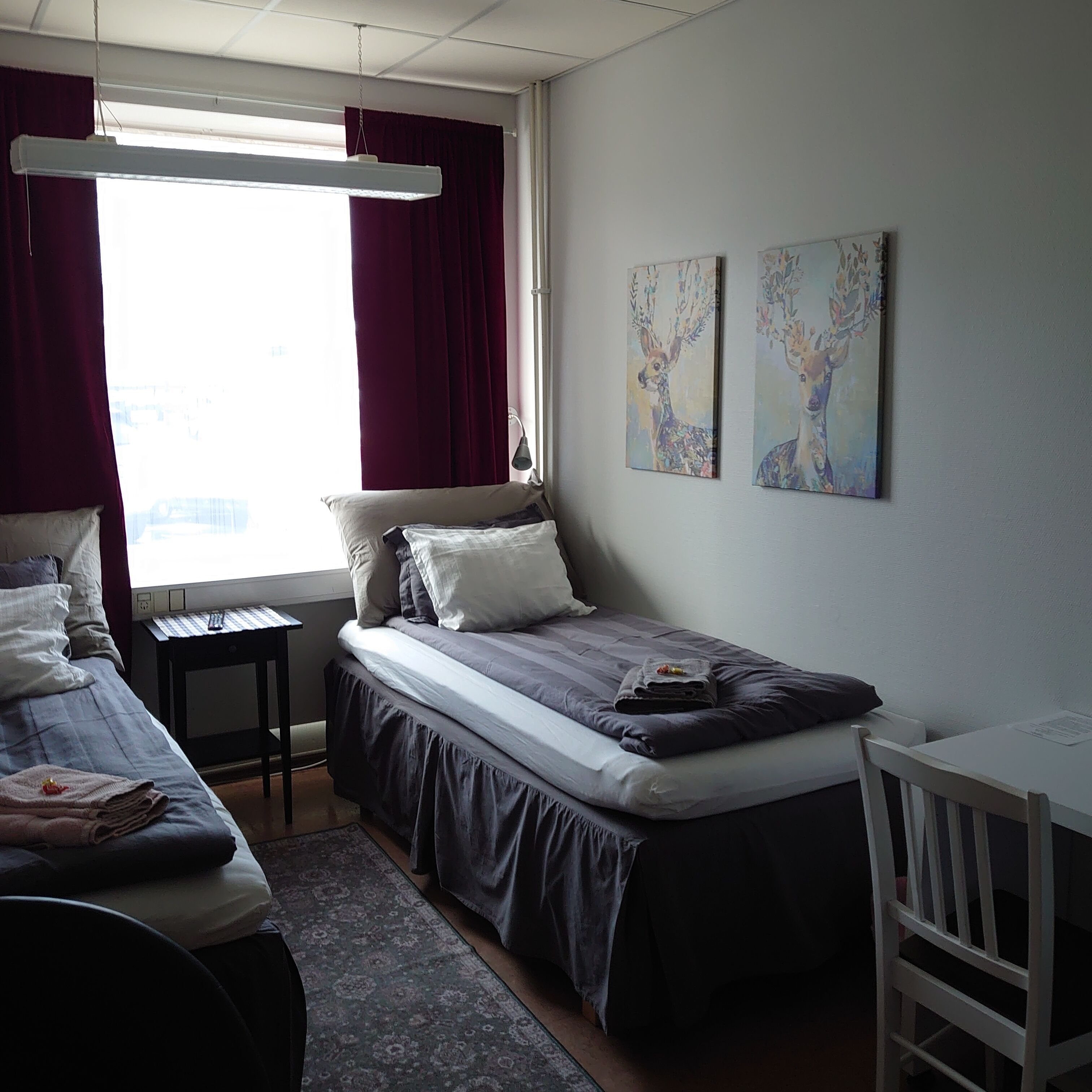 Een kleine, netjes ingerichte kamer met een enkel bed, een bureau en een stoel. Twee schilderijen hangen aan de muur en er is een raam met gordijnen.