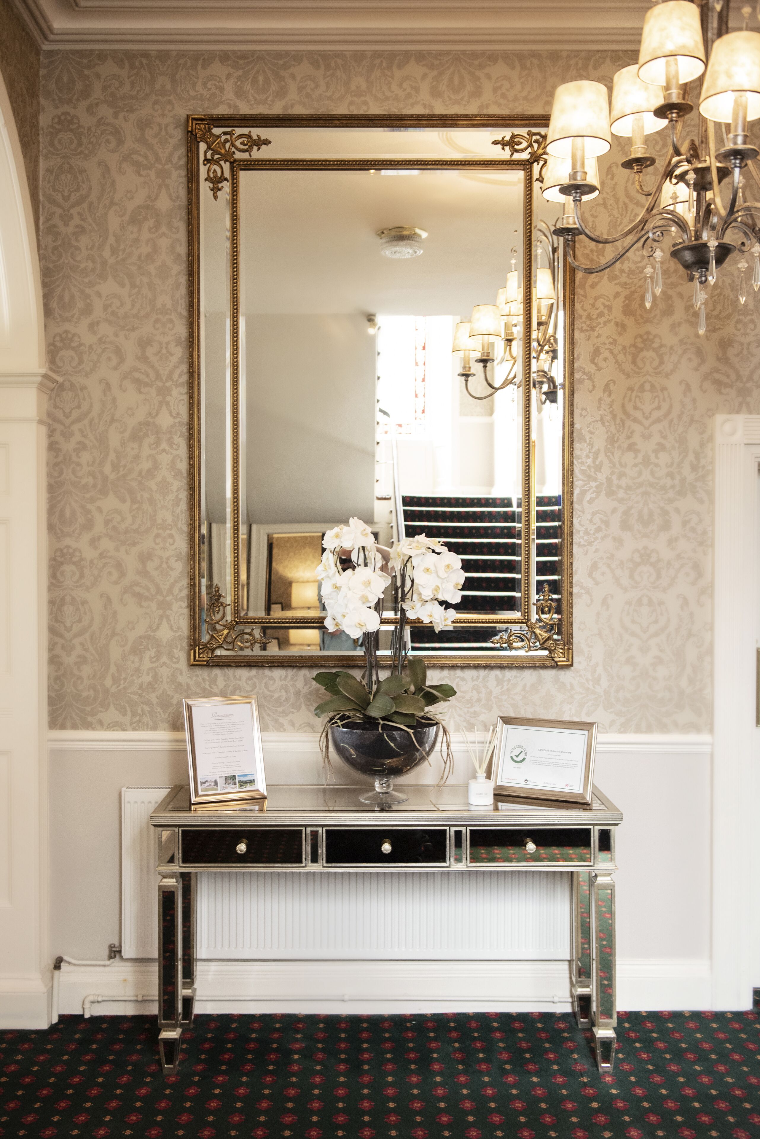 A reception area with a large mirror, a console table with a vase of flowers, and a chandelier.