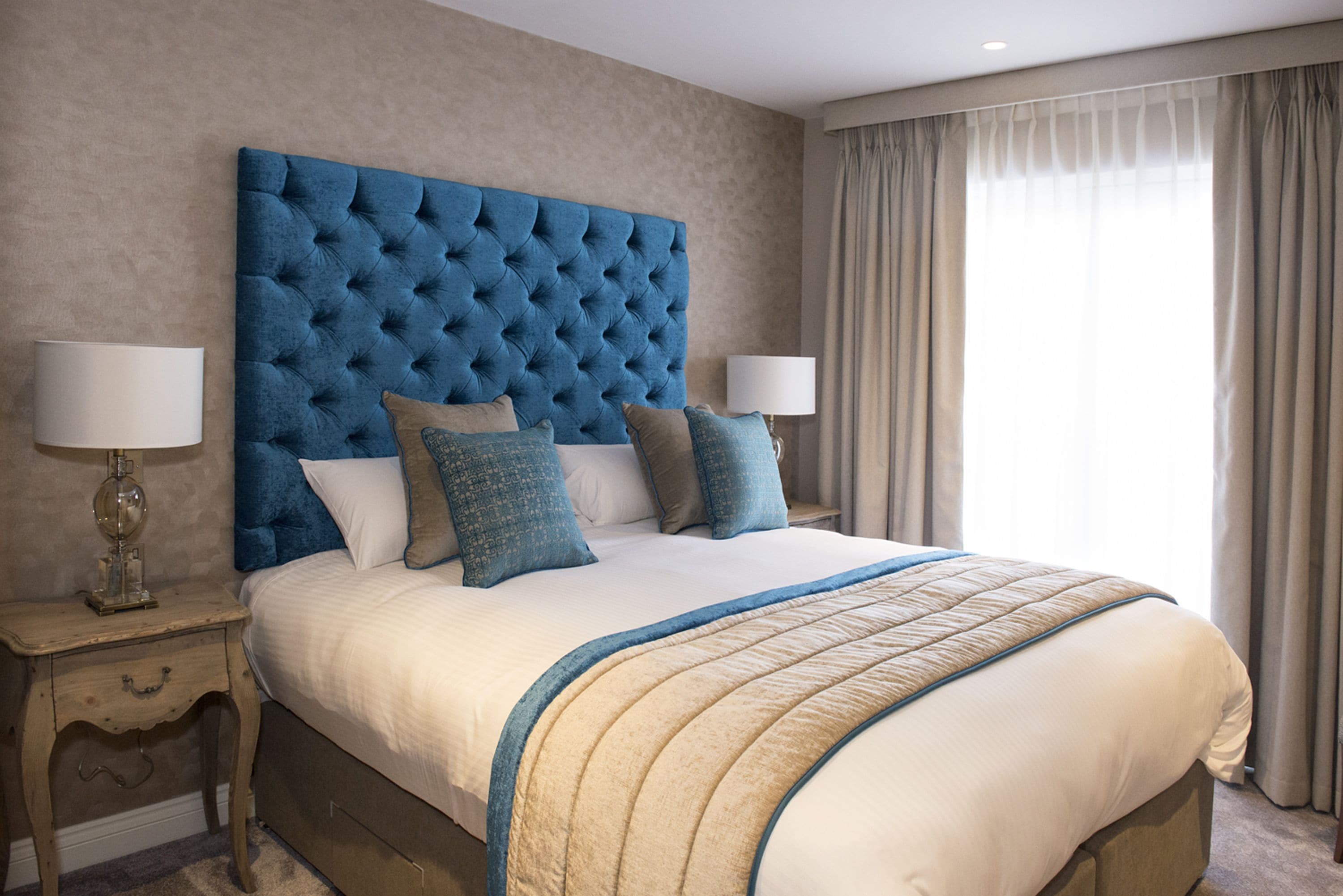 Una cama grande con cabecera capitonada, dos mesitas de noche con lámparas y una ventana con cortinas.