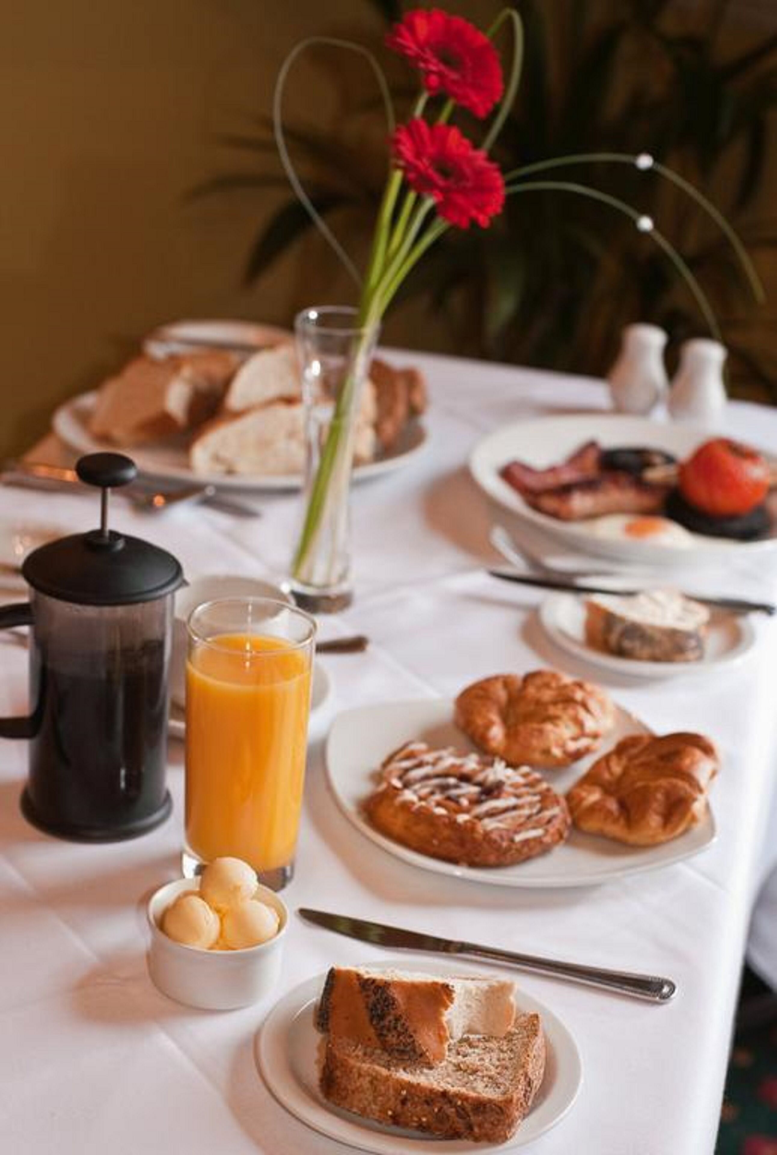 A breakfast table with croissants, a coffee pot, a glass of orange juice, and a vase with red flowers.