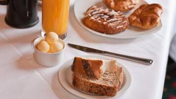 Una mesa de desayuno con cruasanes, una cafetera, un vaso de jugo de naranja y un florero con flores rojas.