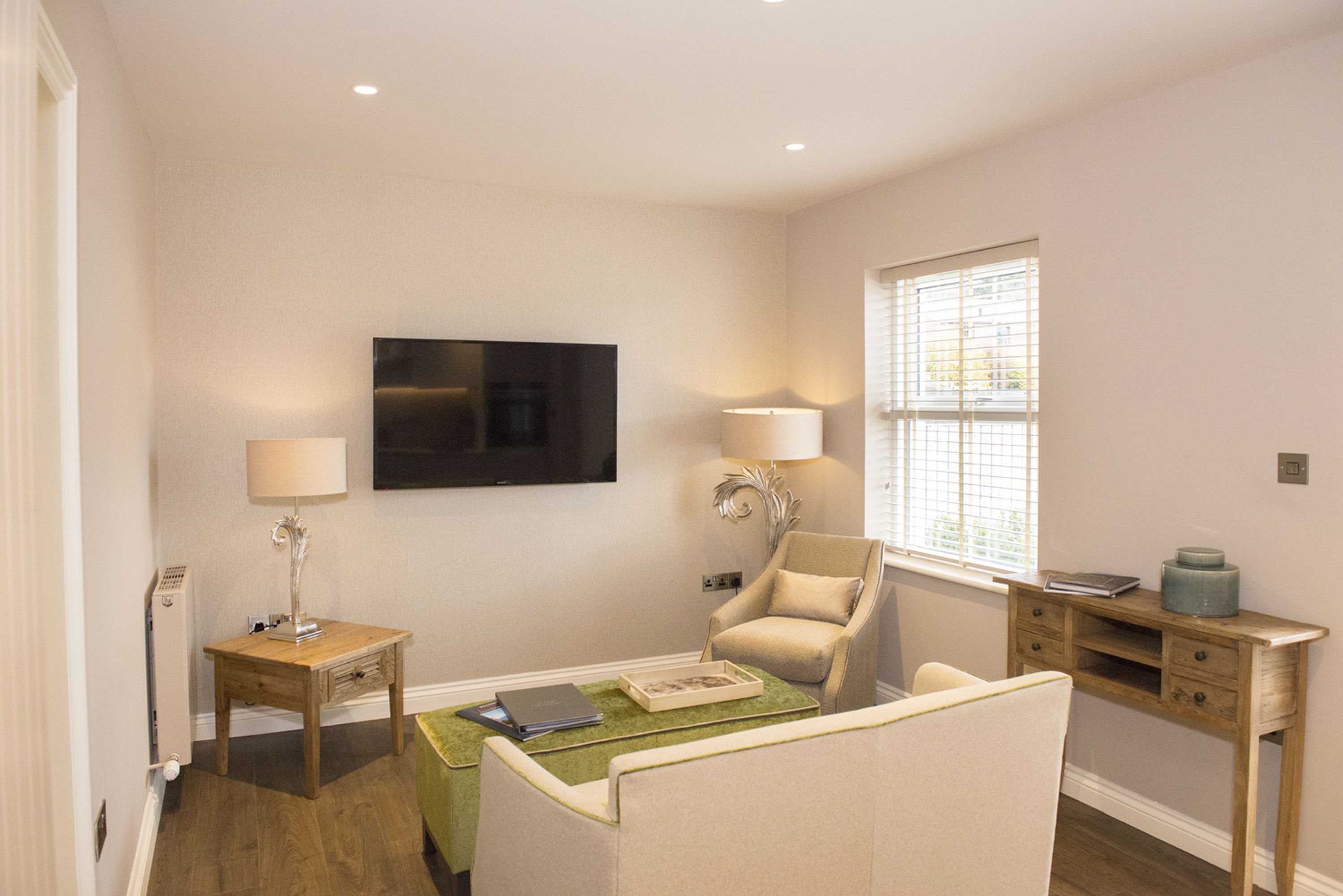 A living room with a flat-screen TV mounted on the wall, a sofa, a chair, a small table with a lamp, and a window with blinds.