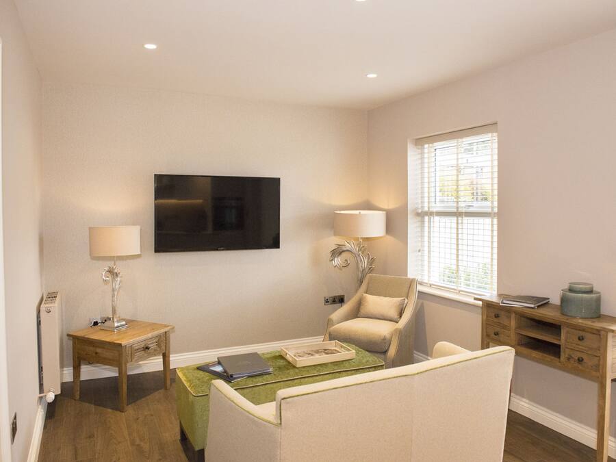A living room with a flat-screen TV mounted on the wall, a sofa, a chair, a small table with a lamp, and a window with blinds.
