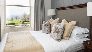 A neatly made bed with a beige bedspread and floral pillows, a bedside lamp, and a view of a green landscape through a large window.