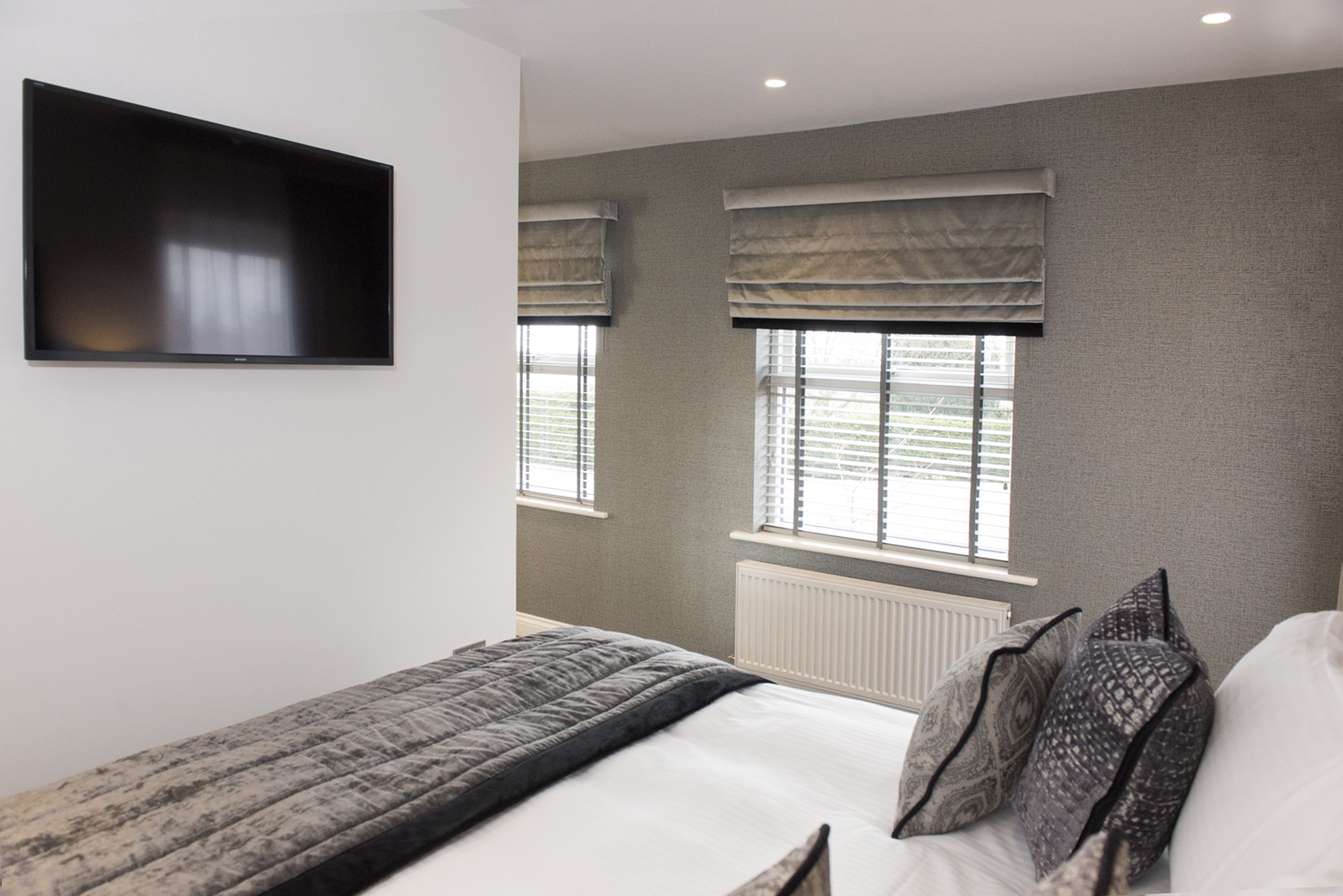 Un dormitorio con cama, televisión de pantalla plana en la pared y dos ventanas con persianas.