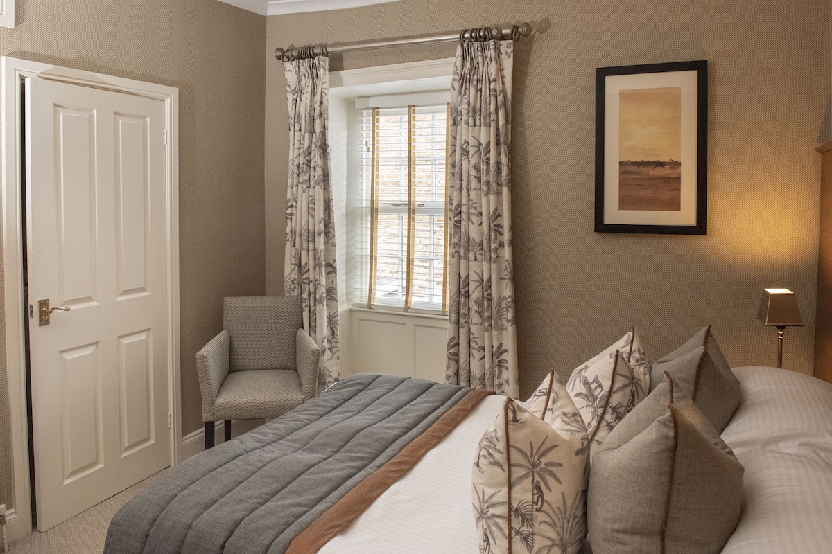 Une chambre à coucher avec un lit, une chaise, une fenêtre avec des rideaux, une porte et un tableau encadré au mur.