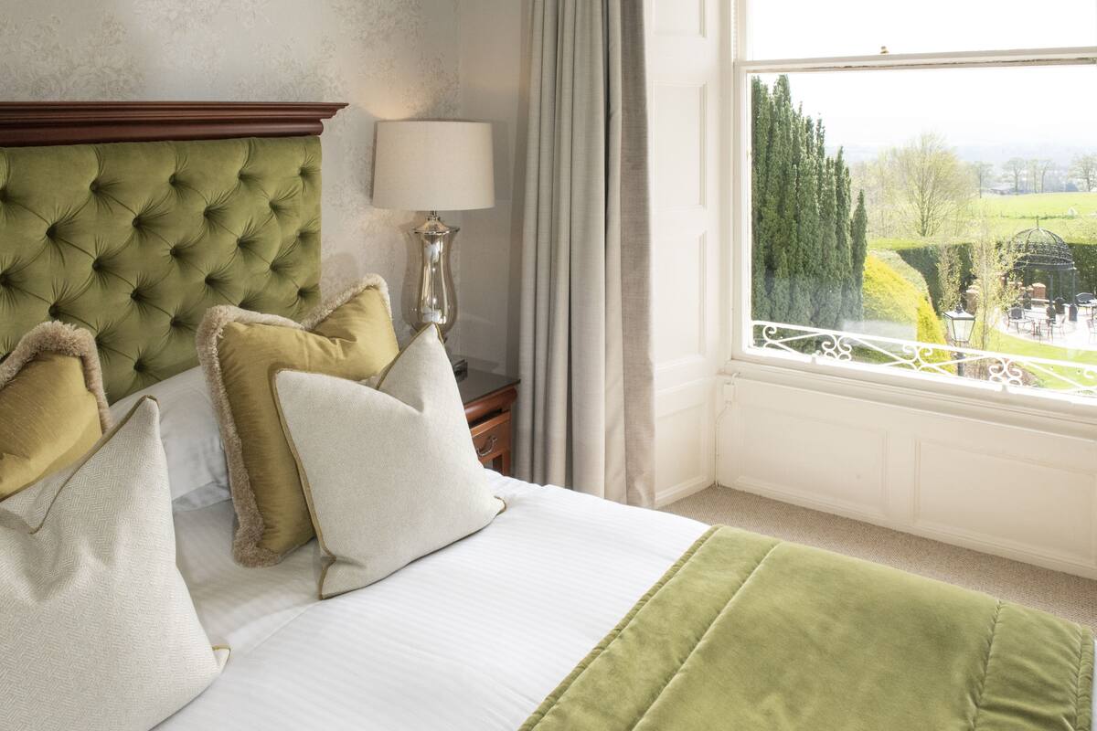 Une chambre d’hôtel avec une tête de lit capitonnée, un lit avec une literie blanche et verte, une lampe et une vue sur un jardin par la fenêtre.