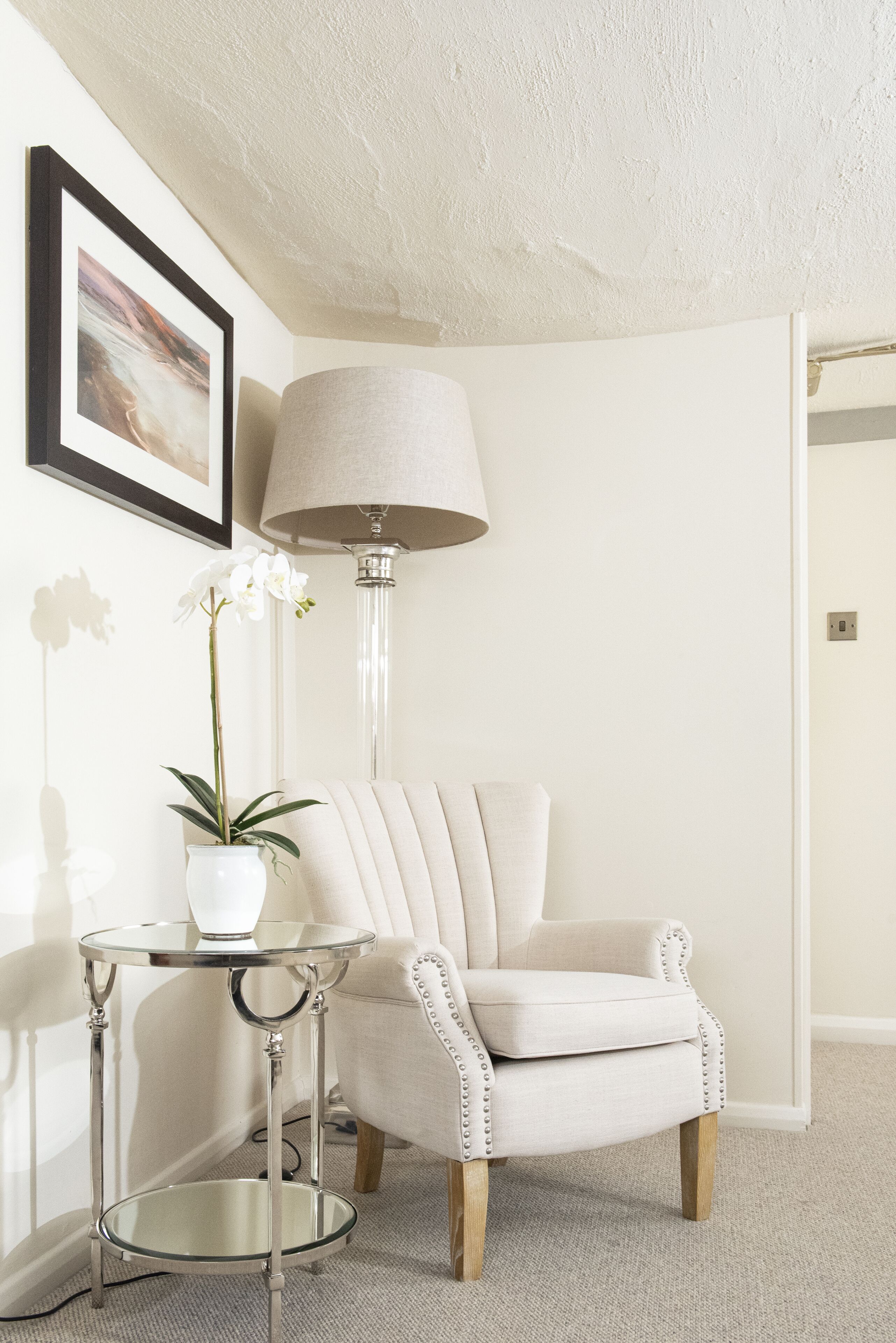 A corner of a room with a white armchair, a small round table with a lamp, and a potted plant.