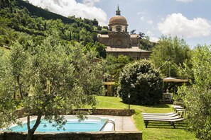 Area piscina con lettini, immersa nel verde e con un edificio storico con cupola sullo sfondo.