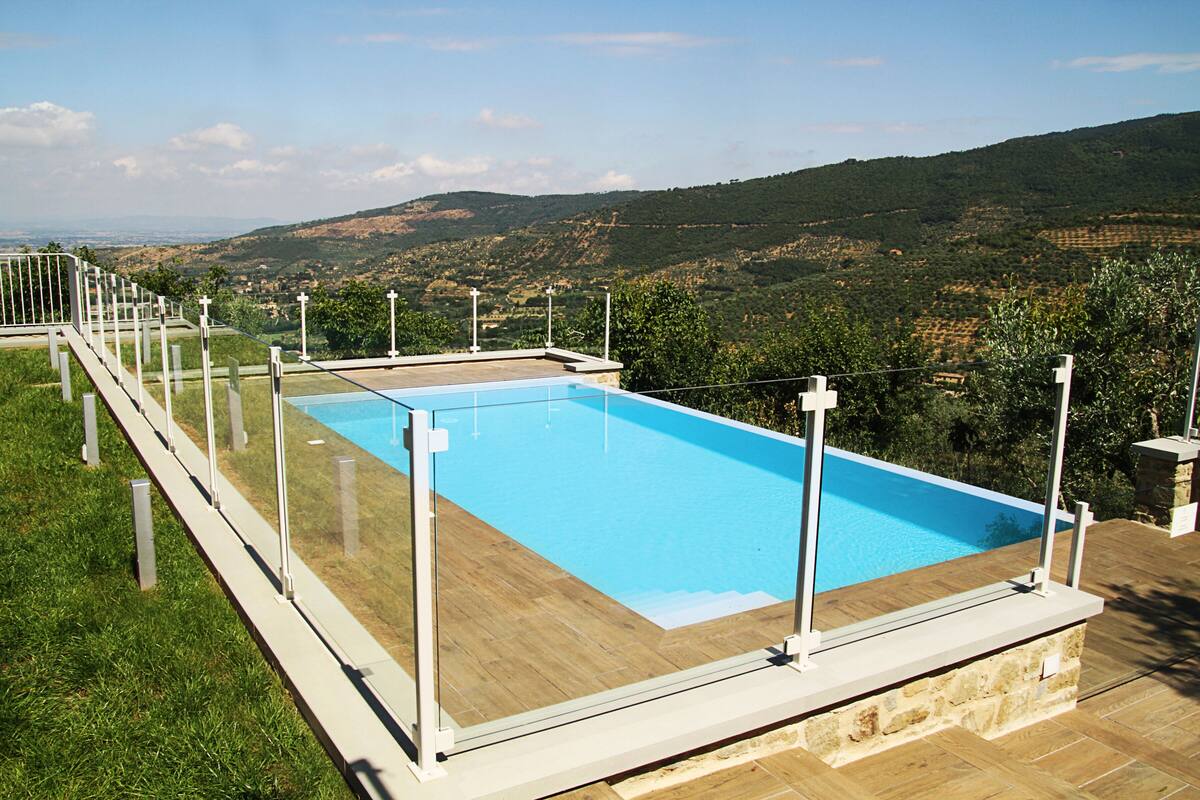 Ein rechteckiger Swimmingpool mit Glasveranda, umgeben von einer Holzterrasse und Steinmauern, vor einer Kulisse aus sanften Hügeln und blauem Himmel.