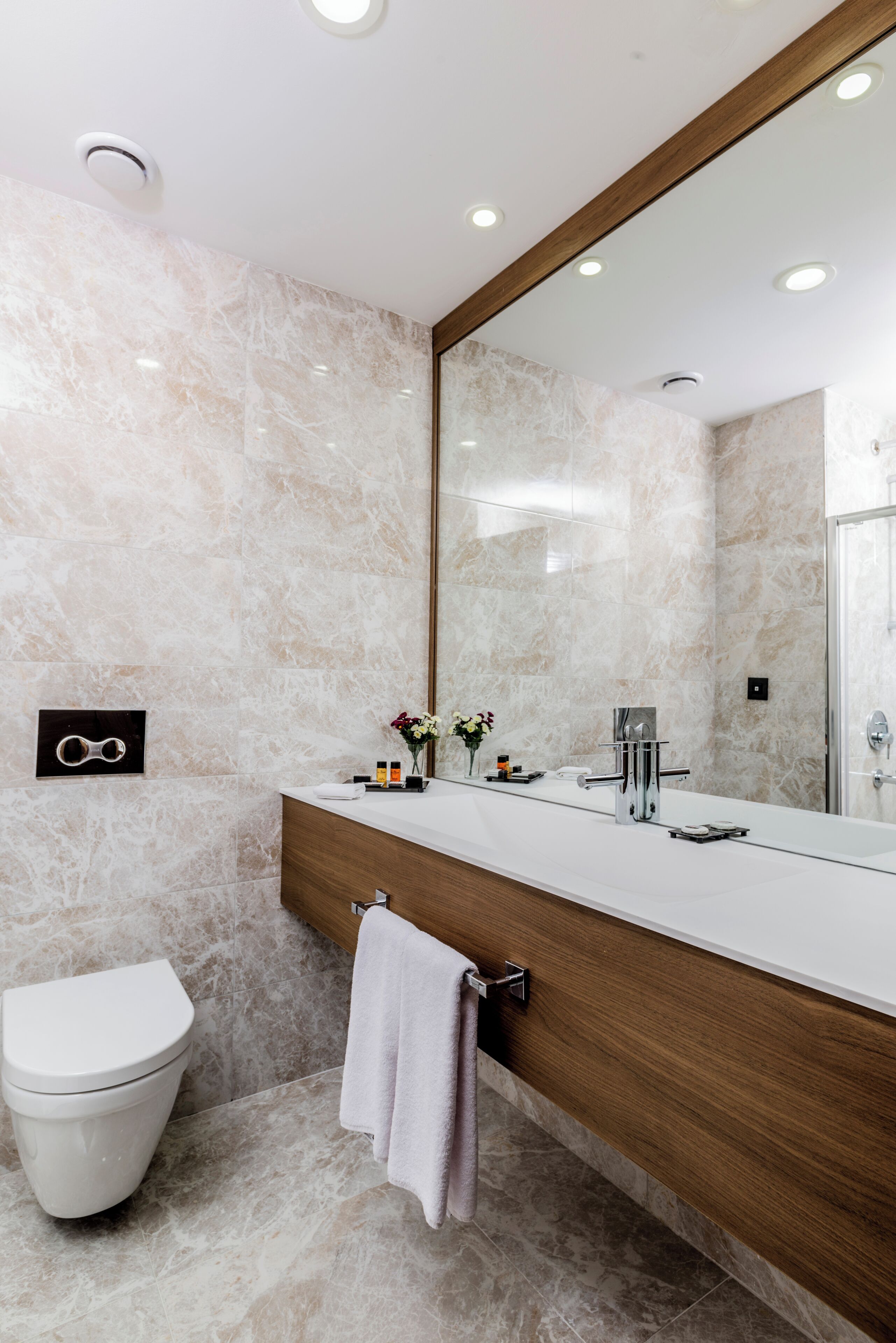 Ein modernes Badezimmer mit Marmorwand, Waschtisch aus Holz, großem Spiegel, Toilette und Handtuchhalter.