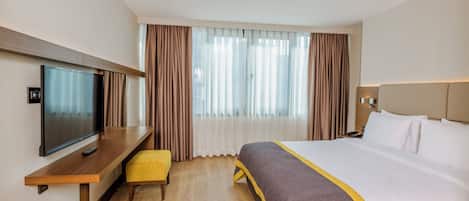 A hotel room with a bed, a TV, a desk, and a window with curtains.