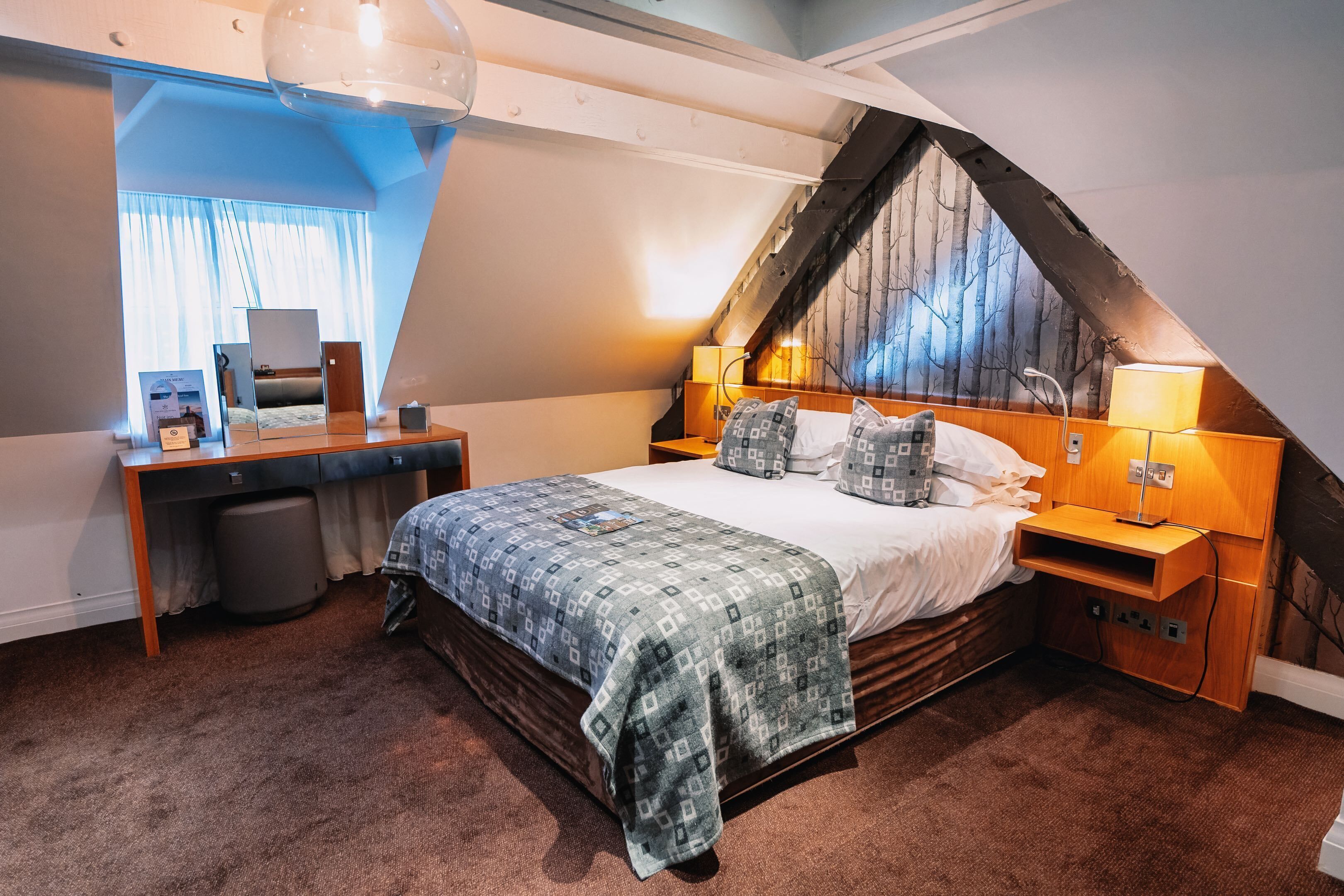 A bedroom with a sloped ceiling, a bed with patterned bedding, a desk with a computer, and a window with curtains.