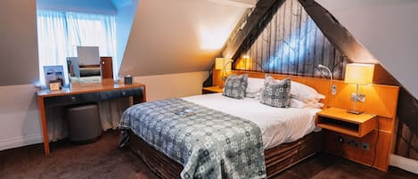 A bedroom with a sloped ceiling, a bed with patterned bedding, a desk with a computer, and a window with curtains.