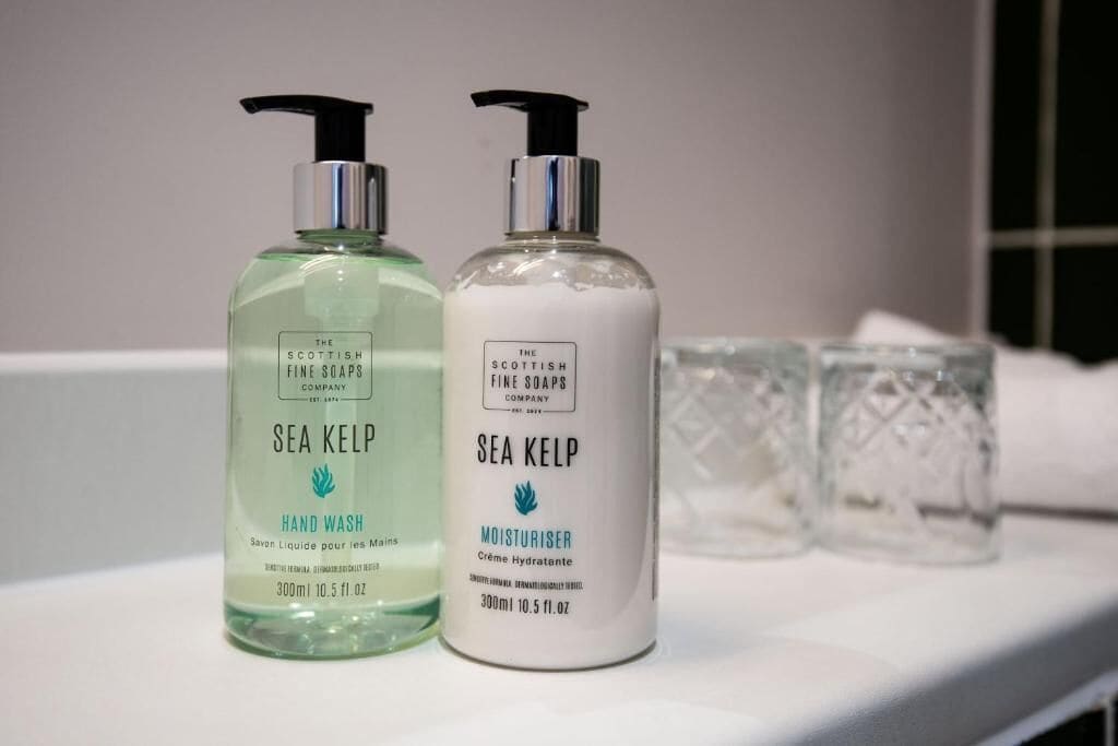 Deux flacons de savon pour les mains et de crème hydratante à base d’algues marines de la marque The Scottish Fine Soaps Company, posés sur une surface blanche, accompagnés de deux verres transparents.