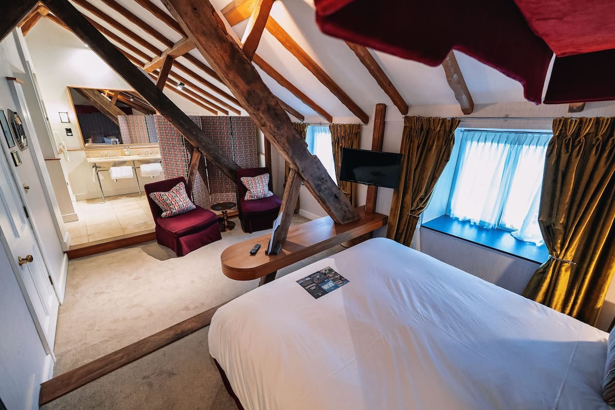 A bedroom with a large bed, a wooden ceiling, a desk with a TV, and a view of the outside.