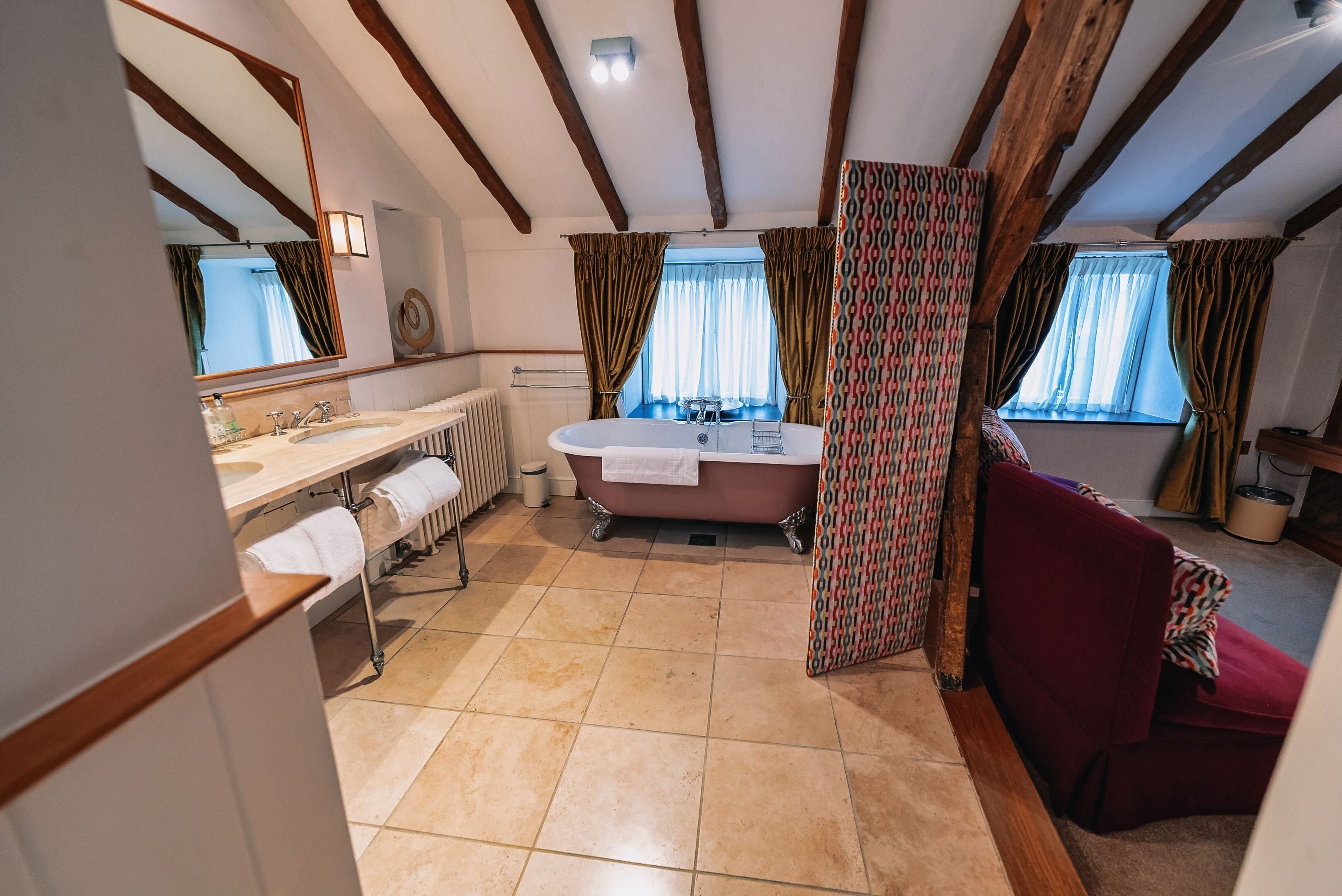 Ein Badezimmer mit einer Badewanne, einem Waschbecken und einem Spiegel. Handtücher hängen an einem Wäscheständer, und ein Fenster mit Vorhängen.