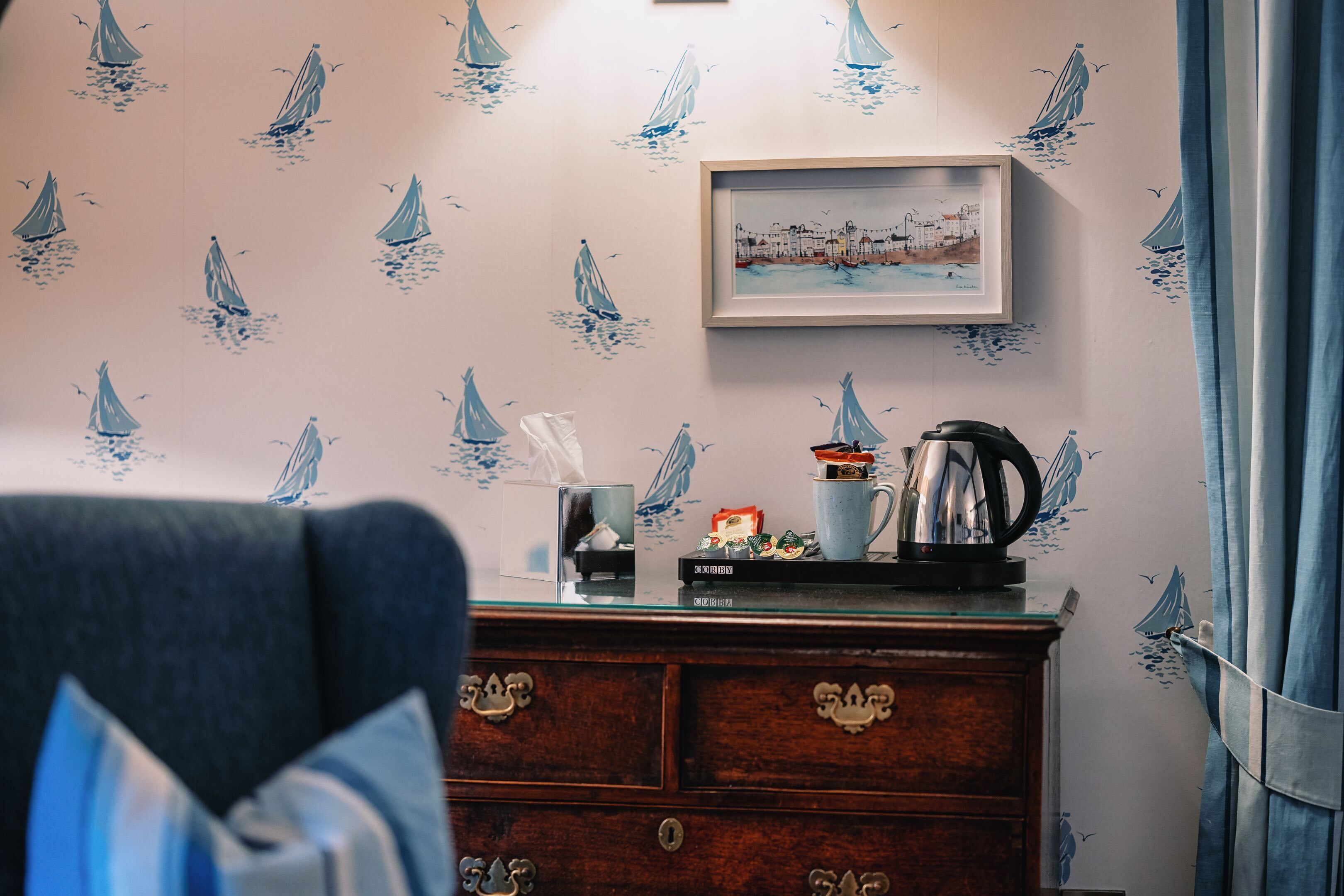 A room with a wall decorated with sailboat patterns, a framed picture, and a wooden dresser with drawers.
