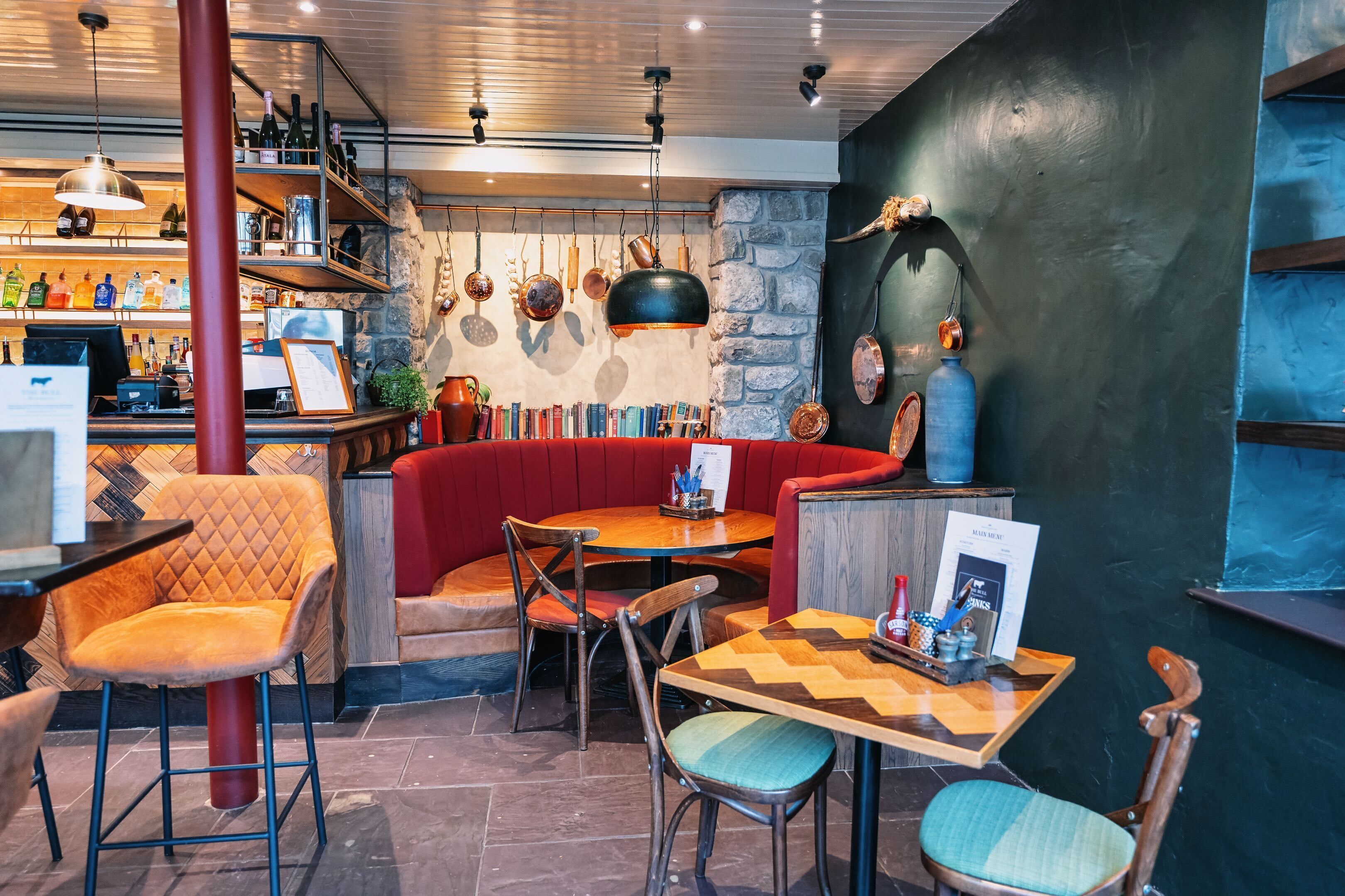 A cozy restaurant with a bar area, cushioned seating, and a variety of bottles and dishes displayed on shelves.