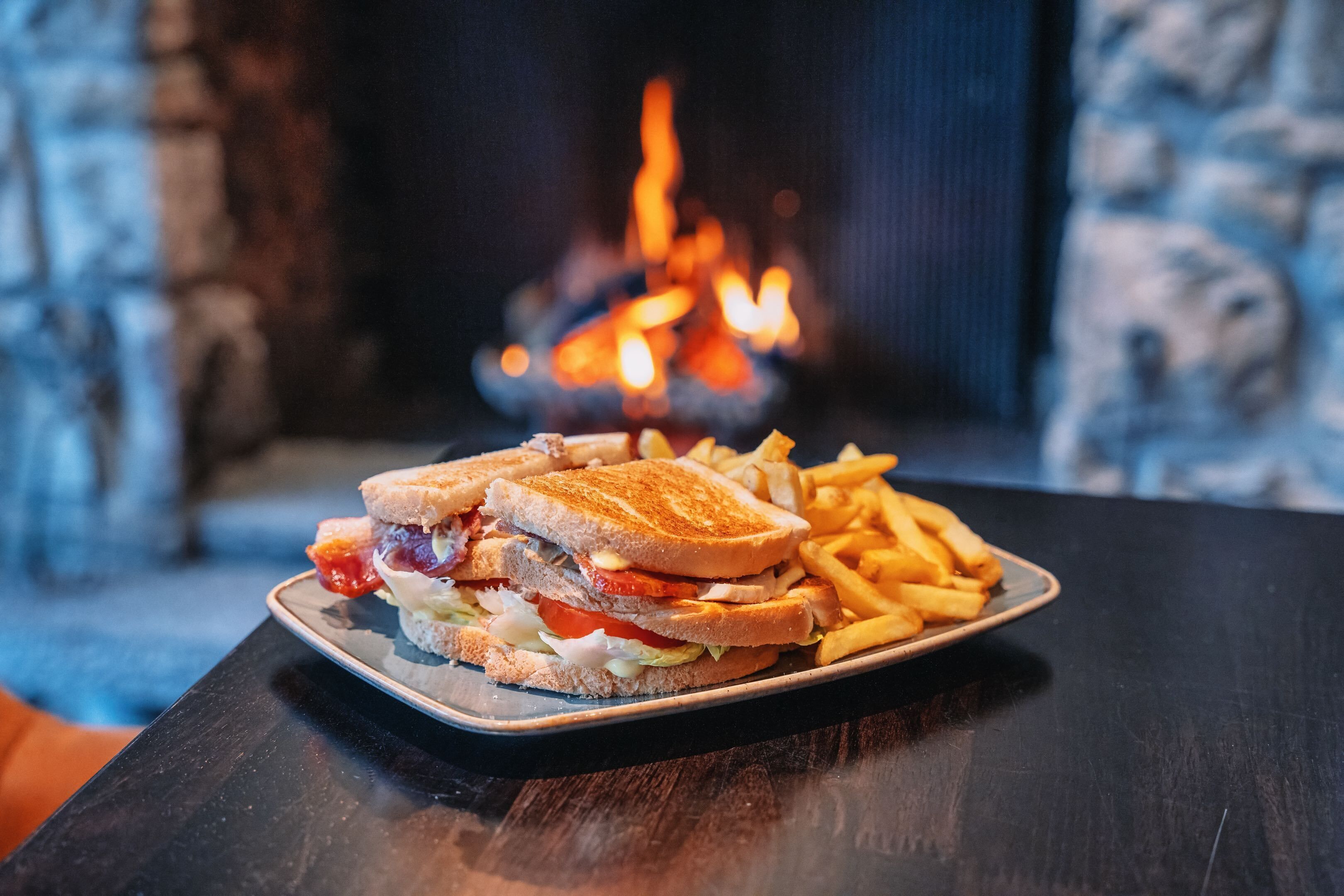 Ein Teller mit einem Sandwich und Pommes auf einem Tisch, im Hintergrund eine Steinwand und ein Kamin.