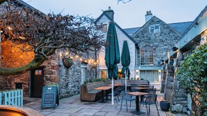 An outdoor patio area with stone walls, wooden benches, and tables. There are green umbrellas and potted plants. - The Bull and Townhouse, Beaumaris- The Inn Collection Group (Beaumaris)