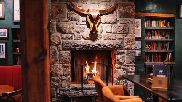 A stone fireplace with a large bull's head above it, a bookshelf to the right, and a red booth in the foreground. - The Bull and Townhouse, Beaumaris- The Inn Collection Group (Beaumaris)