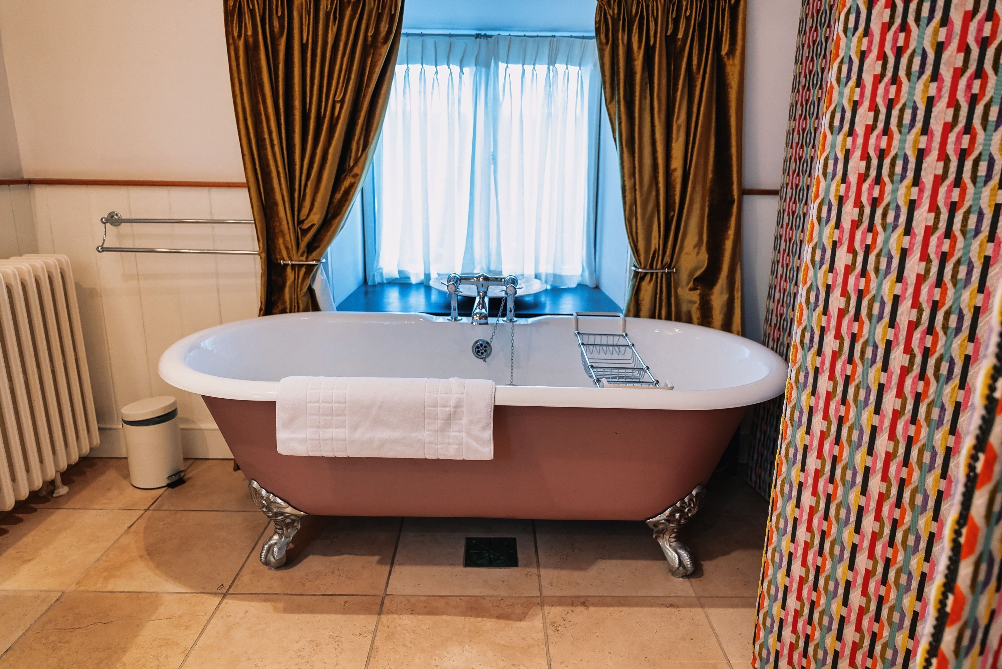 A bathroom with a freestanding bathtub, a window with curtains, a radiator, and a patterned curtain on the wall.