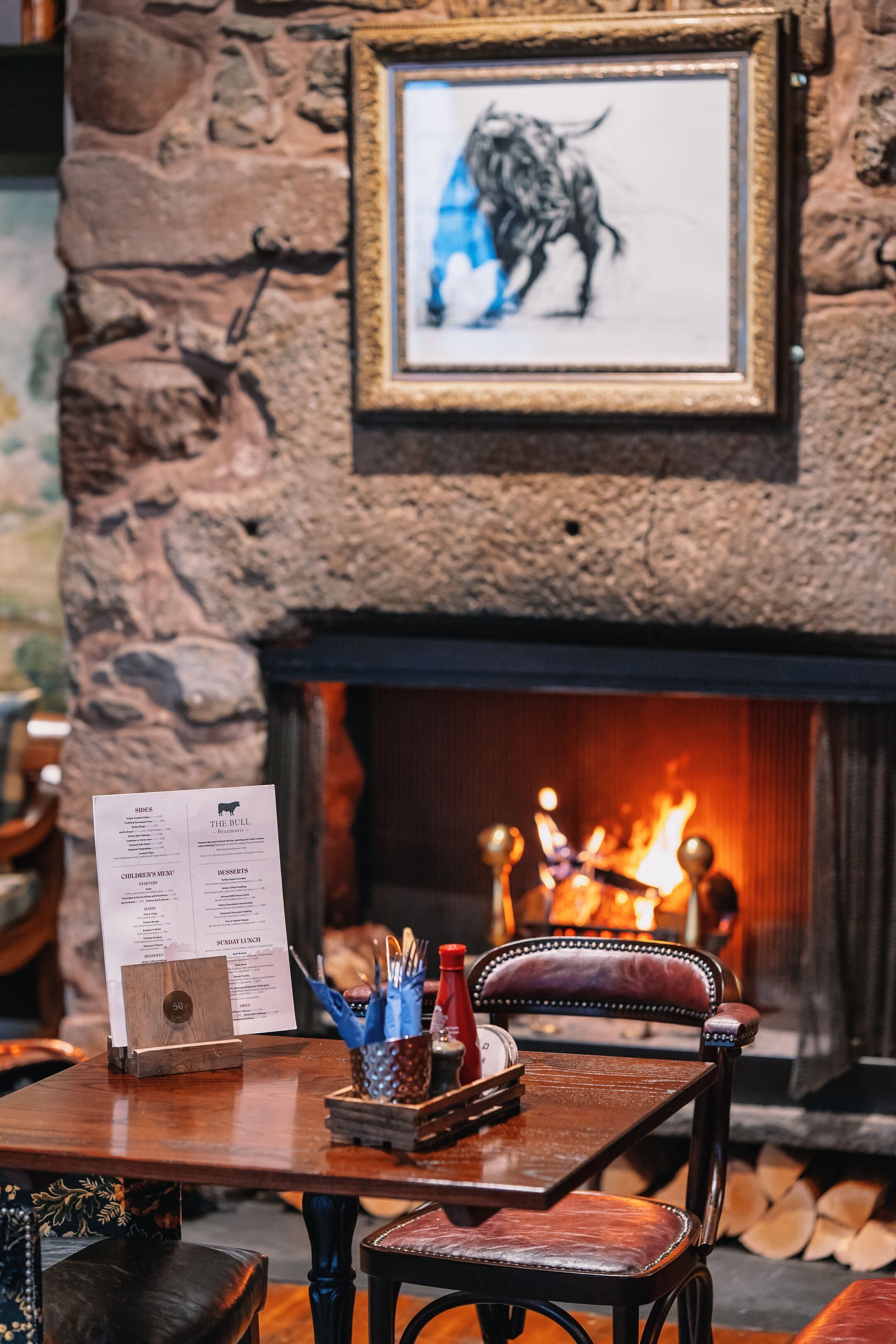 L’intérieur d’un restaurant avec un mur en pierre, un tableau encadré représentant un taureau, un présentoir à menus et une cheminée avec des bûches.