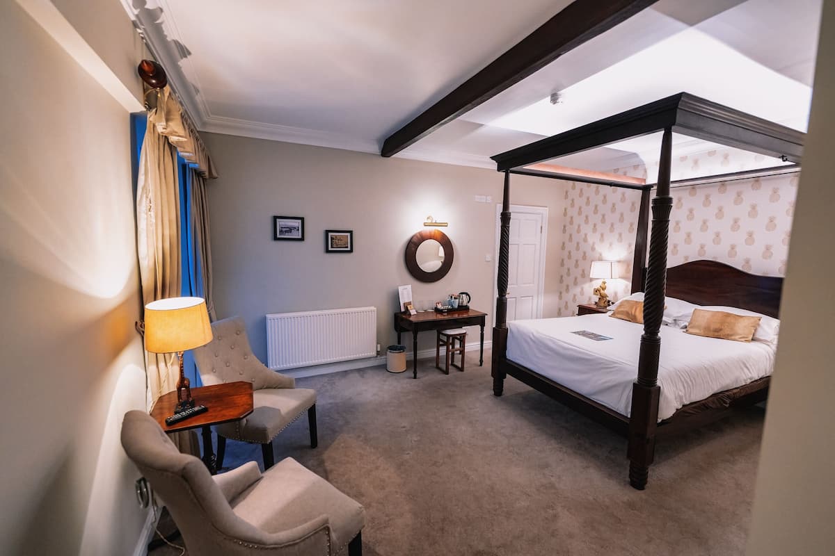 A hotel room with a four-poster bed, a desk with a lamp, a chair, and a small table.
