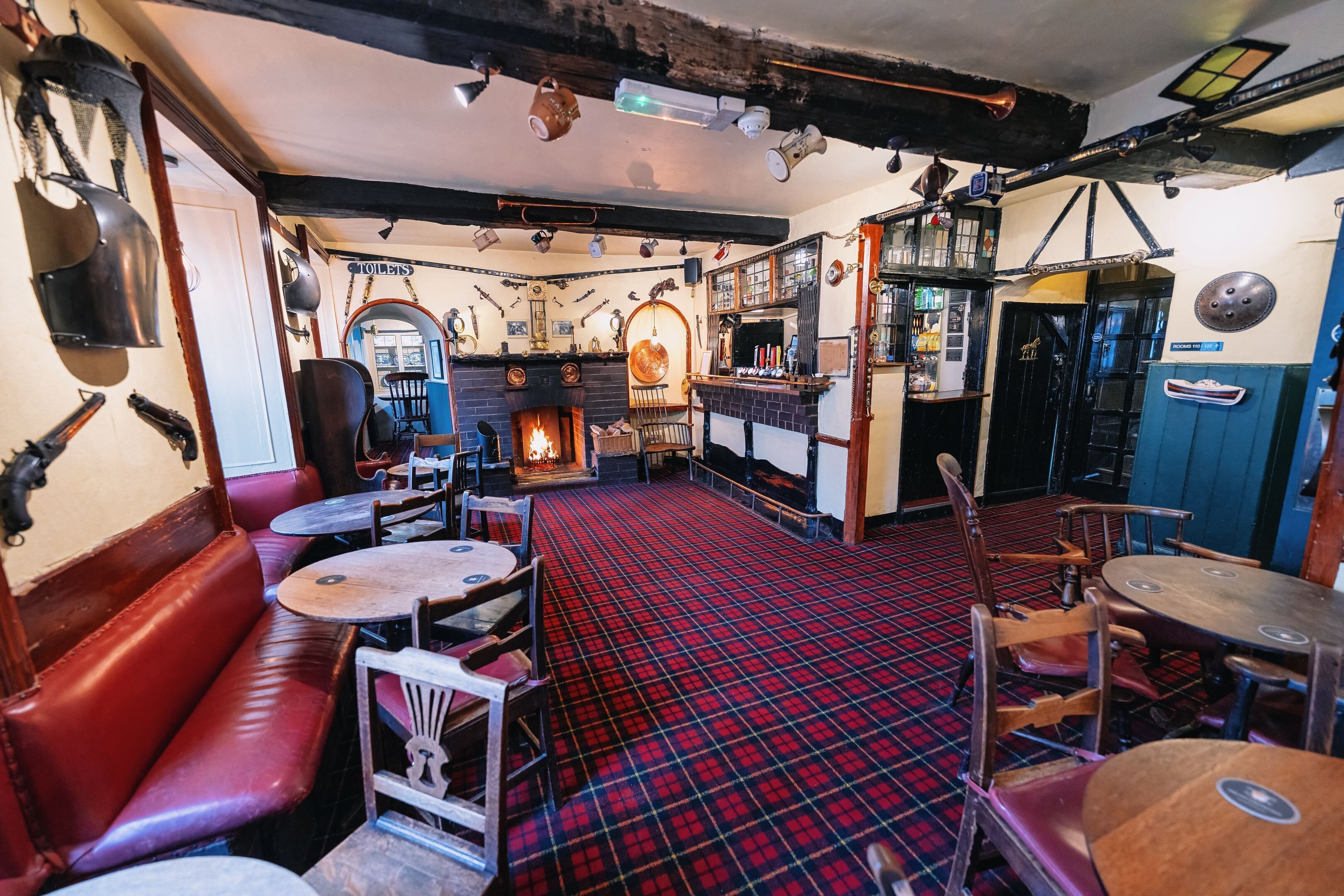 A pub with red plaid flooring, wooden benches, and tables. There is a bar area with stools and a fireplace.