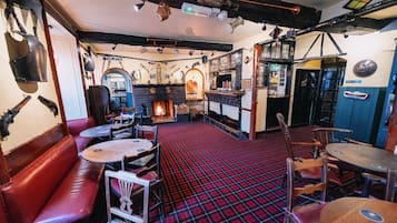 A pub with red plaid flooring, wooden benches, and tables. There is a bar area with stools and a fireplace.