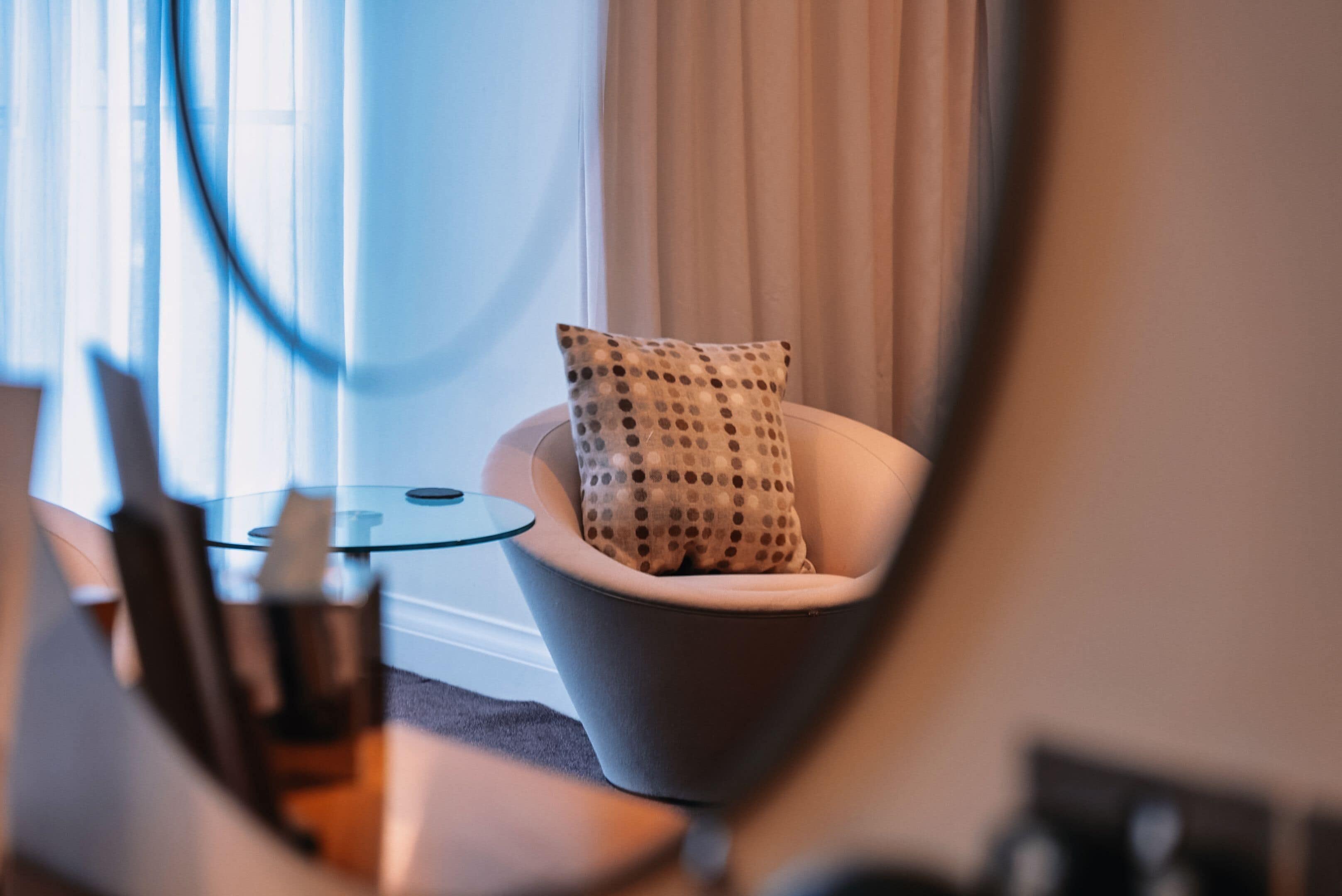 Una habitación moderna con una mesa de vidrio, una silla blanca con un cojín de lunares y un espejo grande.