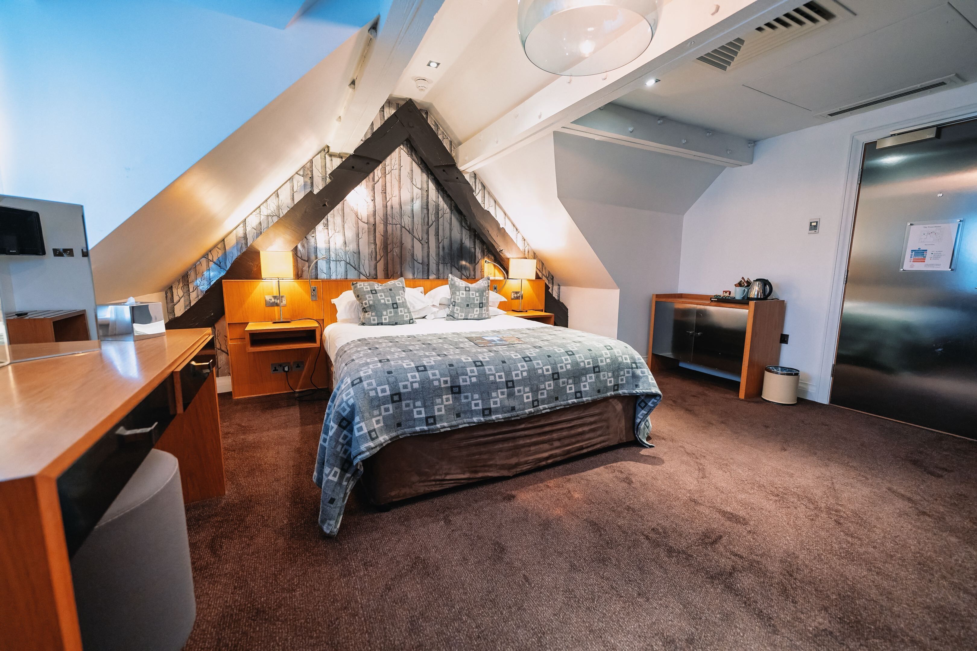 Une chambre avec un plafond en pente, un lit avec une literie à motifs, un bureau et un réfrigérateur.