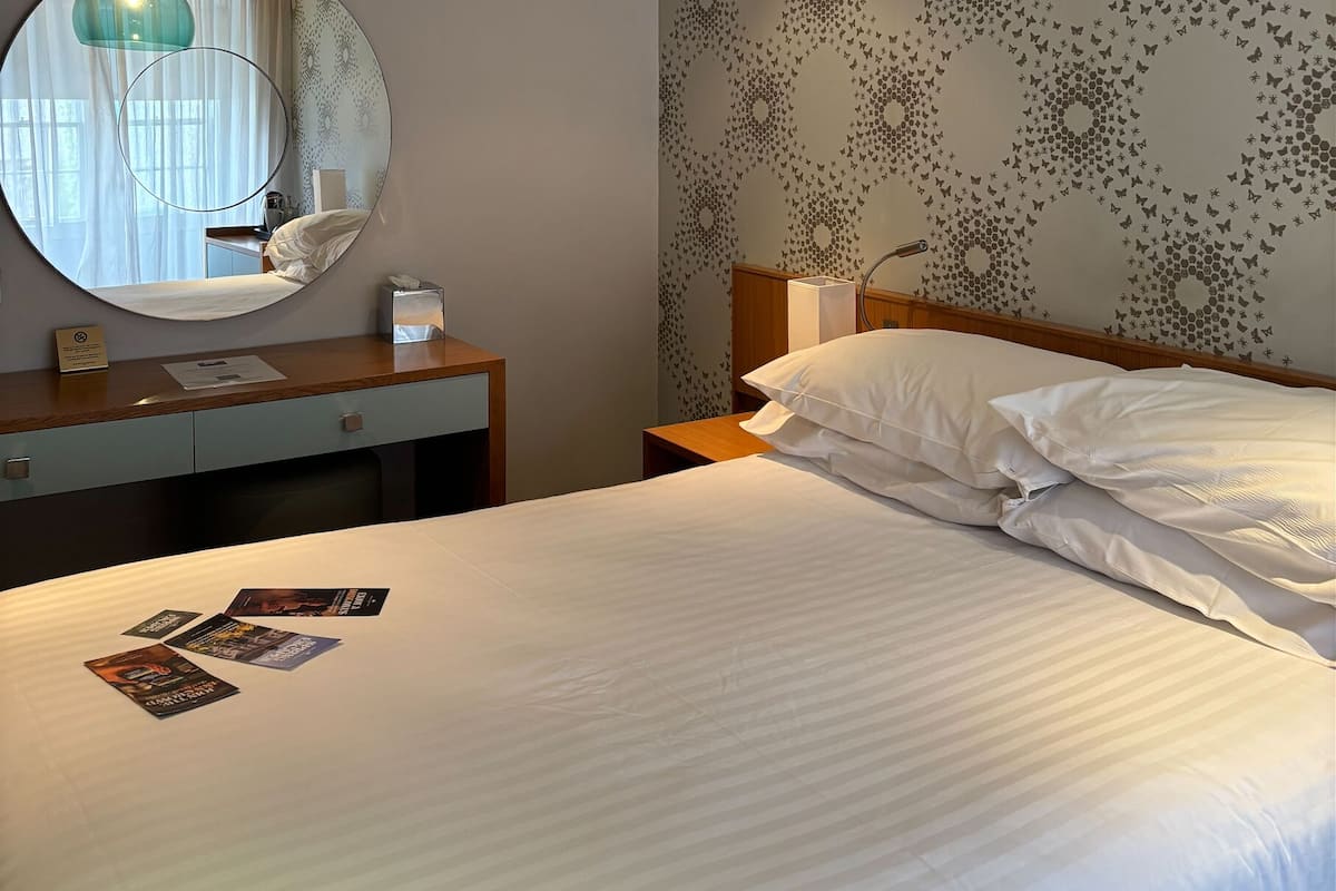 A hotel room with a bed, a round mirror, a bedside table, a lamp, and a wall with a butterfly pattern.