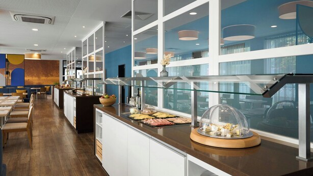 A buffet area with a variety of food items, a glass display case, and a seating area with tables and chairs. - Four Points Flex by Sheraton Essen (Essen)