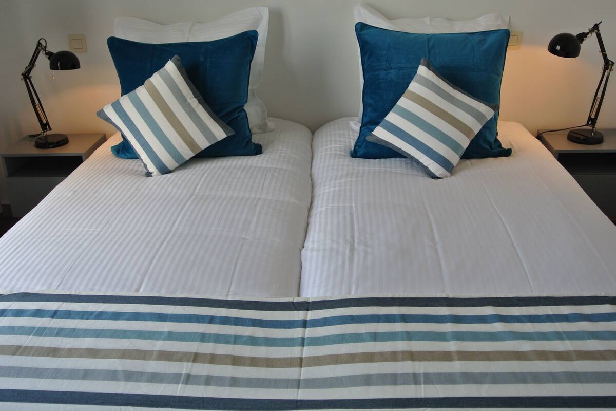 Cama doble con una colcha a rayas y dos almohadas, una a rayas y otra azul lisa, en una habitación de hotel.