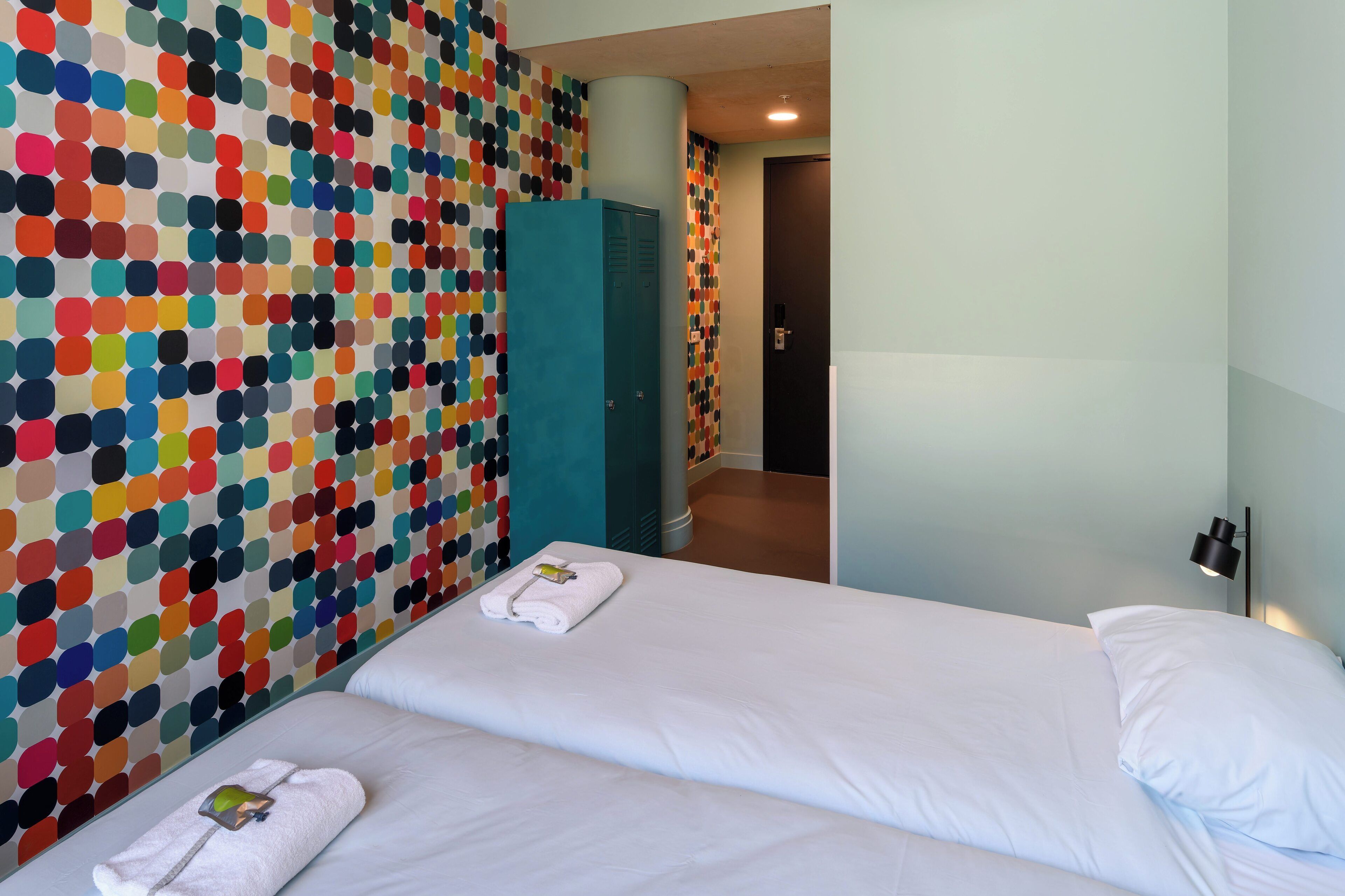 Habitación de hotel con dos camas, un pared con un estampado de lunares y un armario color turquesa, y una puerta al fondo.